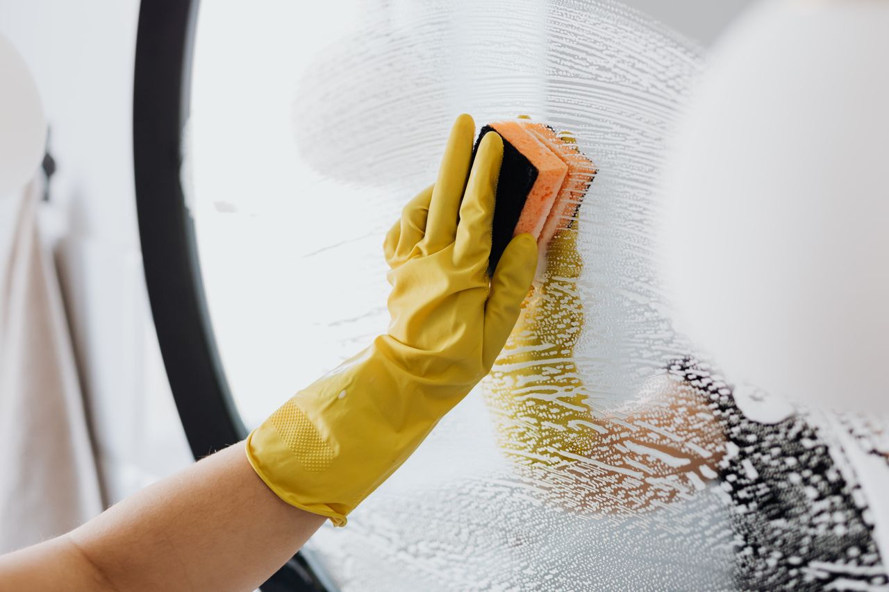 Personne anonyme portant des gants en caoutchouc jaunes et utilisant une simple éponge pour nettoyer le miroir dans la salle de bain